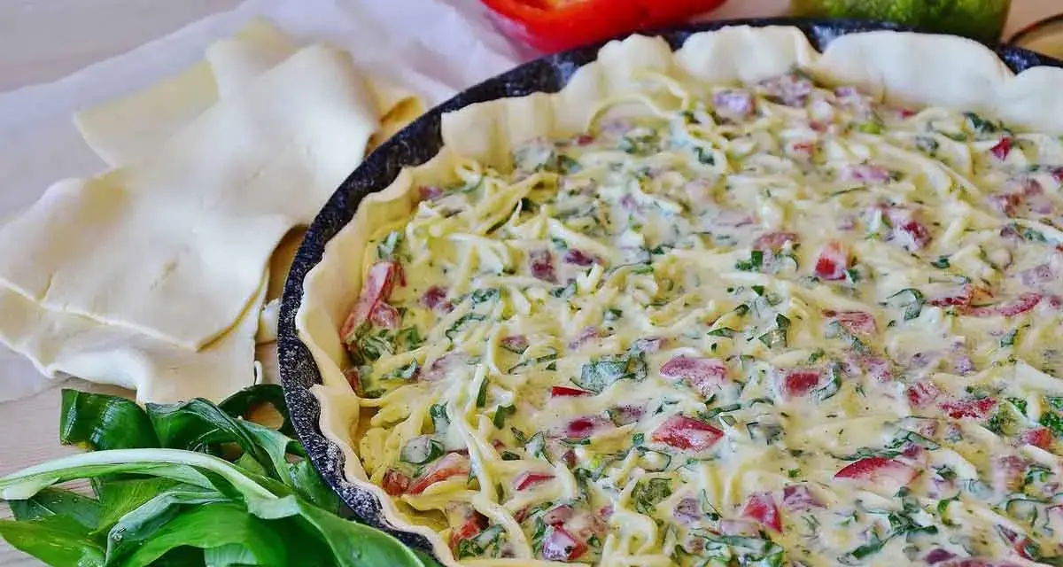 Tarte salée garnie de fromage, poivrons et ail des ours, prête à cuire. Pâte feuilletée et poivron rouge à côté.