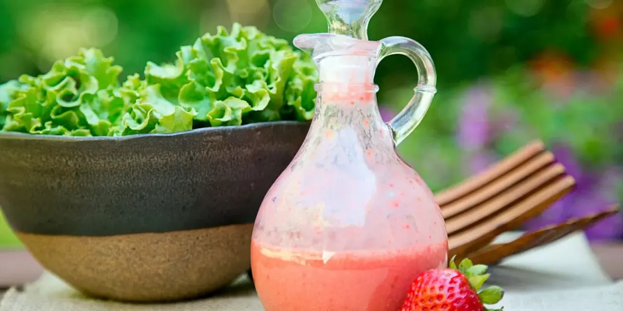 Erdbeeren mit Balsamico-Dressing in einer Karaffe, daneben eine Schüssel Salat und eine einzelne Erdbeere.