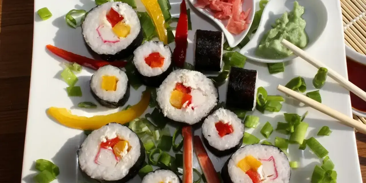 Kolorowe rolki sushi z surimi, papryką i mango, ozdobione szczypiorkiem i papryczkami chili. Obok wasabi i marynowany imbir.