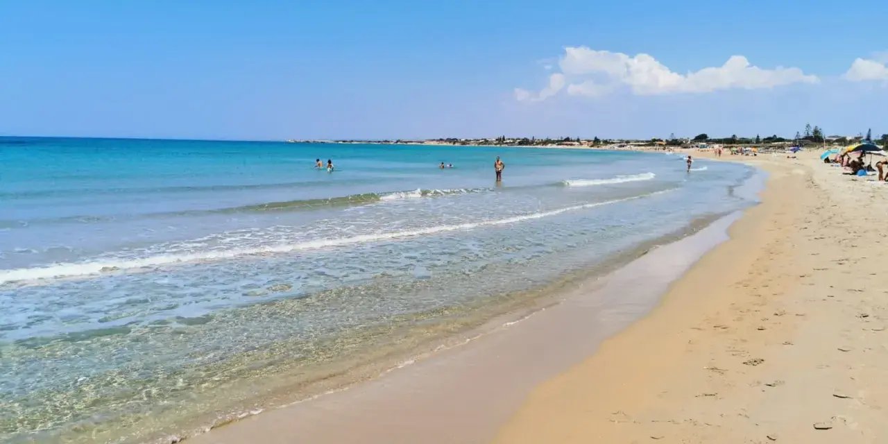 A wide, sandy beach with clear turquoise water and a few people swimming and sunbathing under a bright blue sky.
