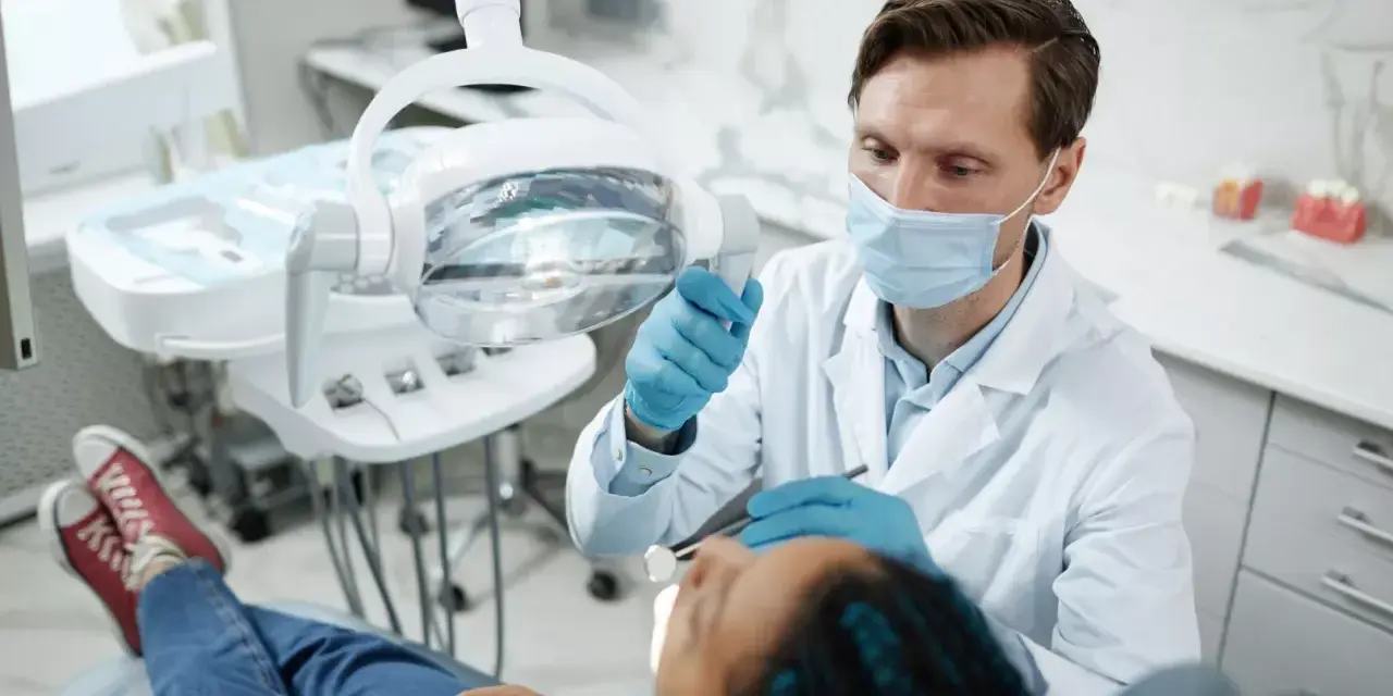 Dentysta w masce i rękawiczkach bada pacjenta w gabinecie stomatologicznym.