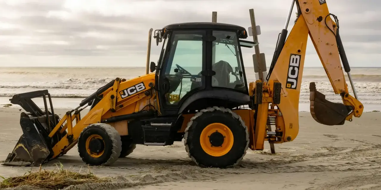 Żółta koparko-ładowarka firmy JCB stoi na piaszczystej plaży, gotowa do pracy.