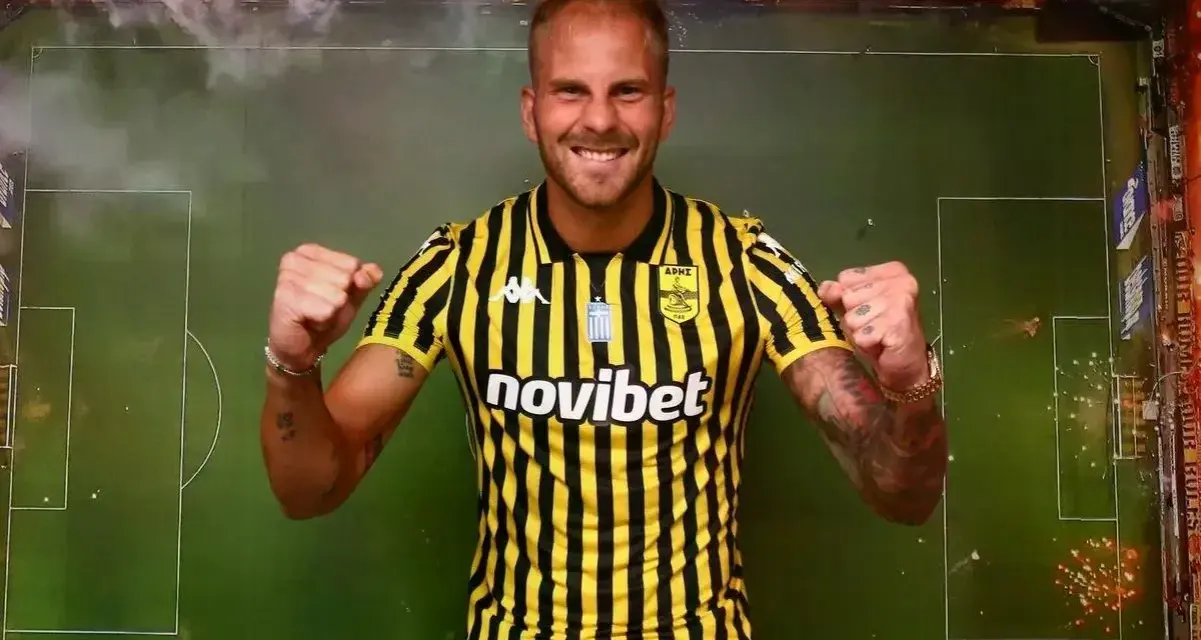 Uros Racic, con su nueva camiseta a rayas amarillas y negras, celebra con los puños en alto. El fondo simula un campo de fútbol con bengalas.