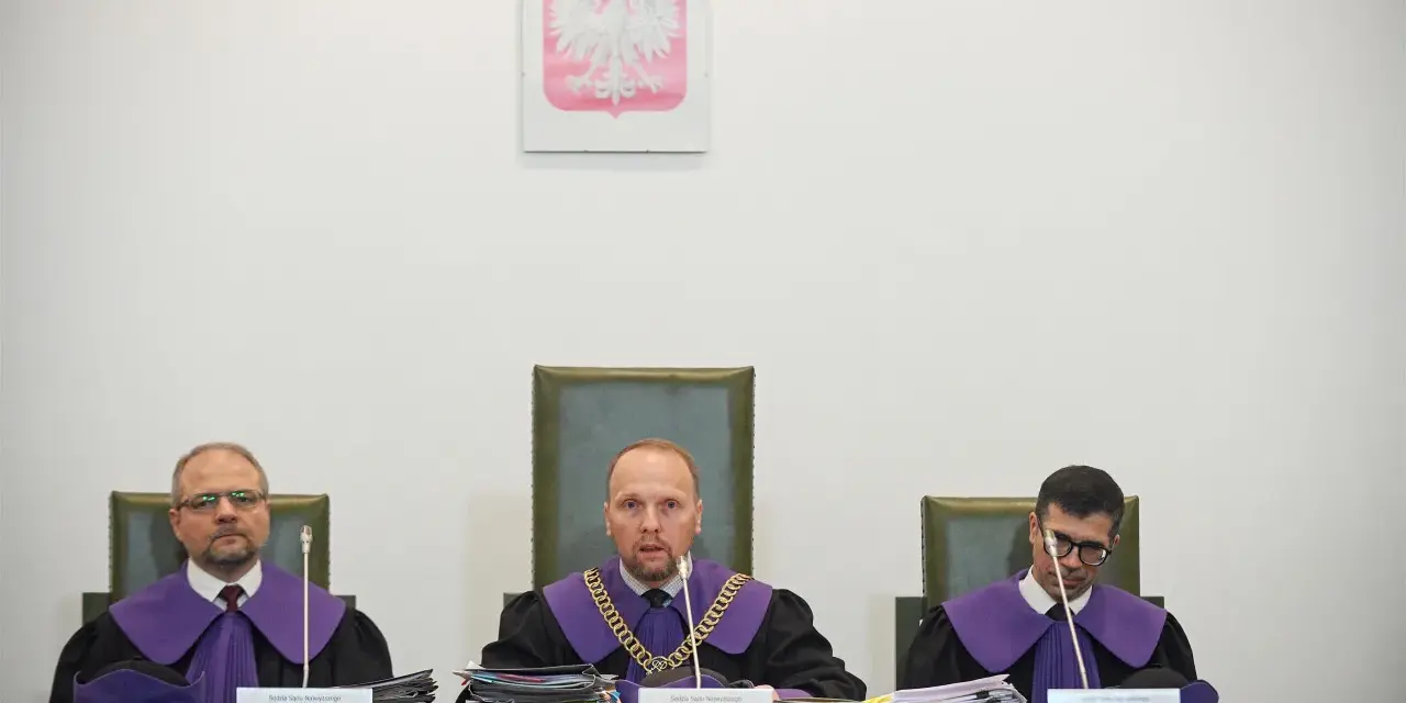 Trzech sędziów w togach podczas jawnego posiedzenia sądu. Na ścianie wisi godło Polski.