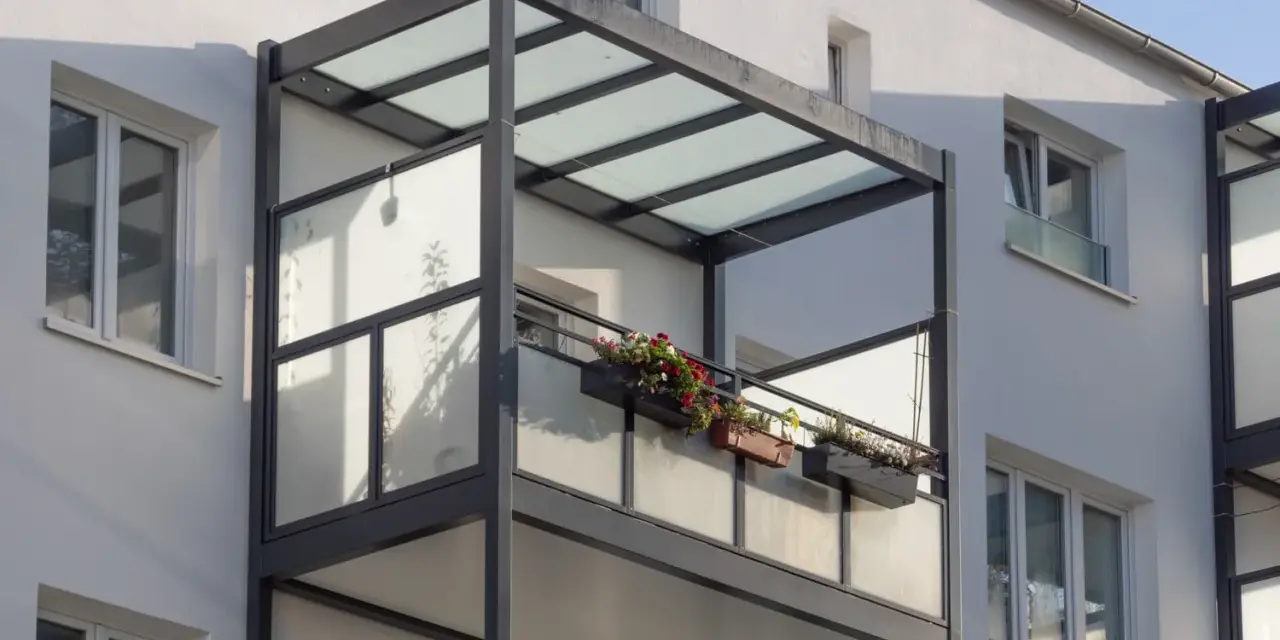 Nowoczesny balkon dostawny z przeszklonym zadaszeniem i balustradą, ozdobiony skrzynkami z kwiatami. Sprawdź balkon dostawny cena.