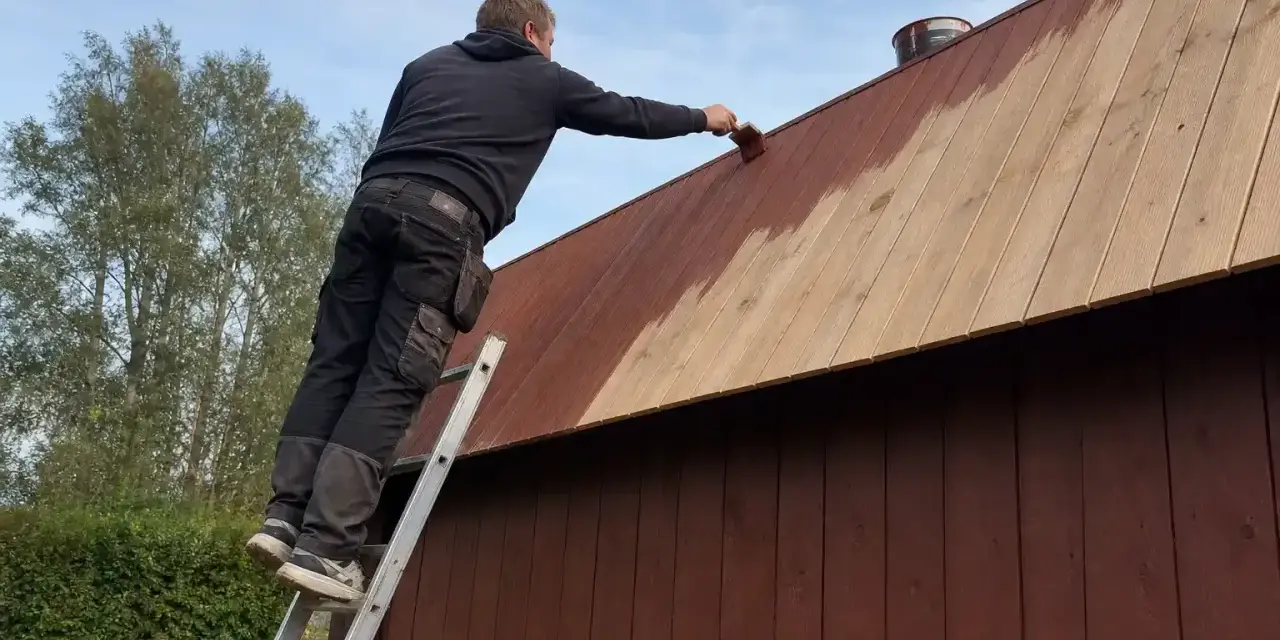 Man på en stege målar plank på ett tak med brun färg.
