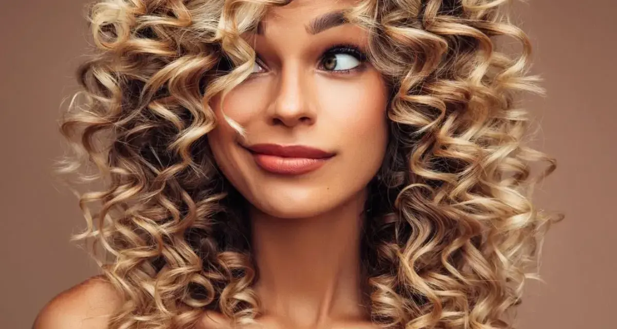 Kobieta z burzą blond loków, krótkie kręcone włosy podkreślają jej uśmiech i spojrzenie.