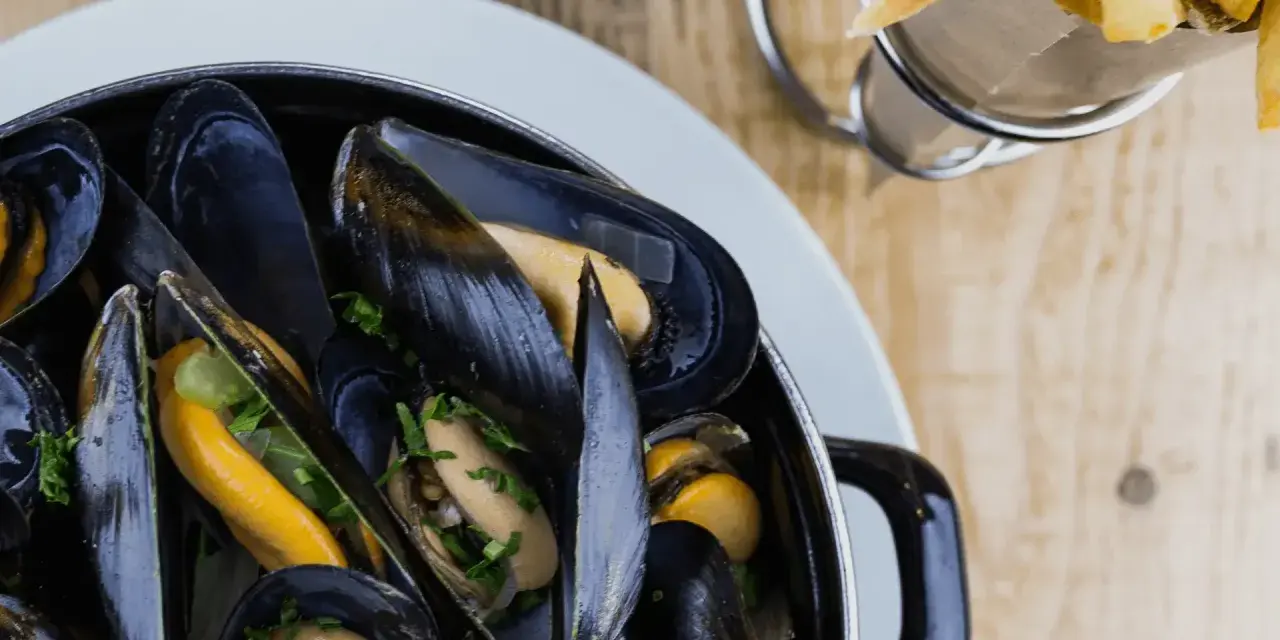 En stor kastrull med ångande blåmusslor, perfekt för en måltid med vin till moules frites. Pommes frites syns i bakgrunden.