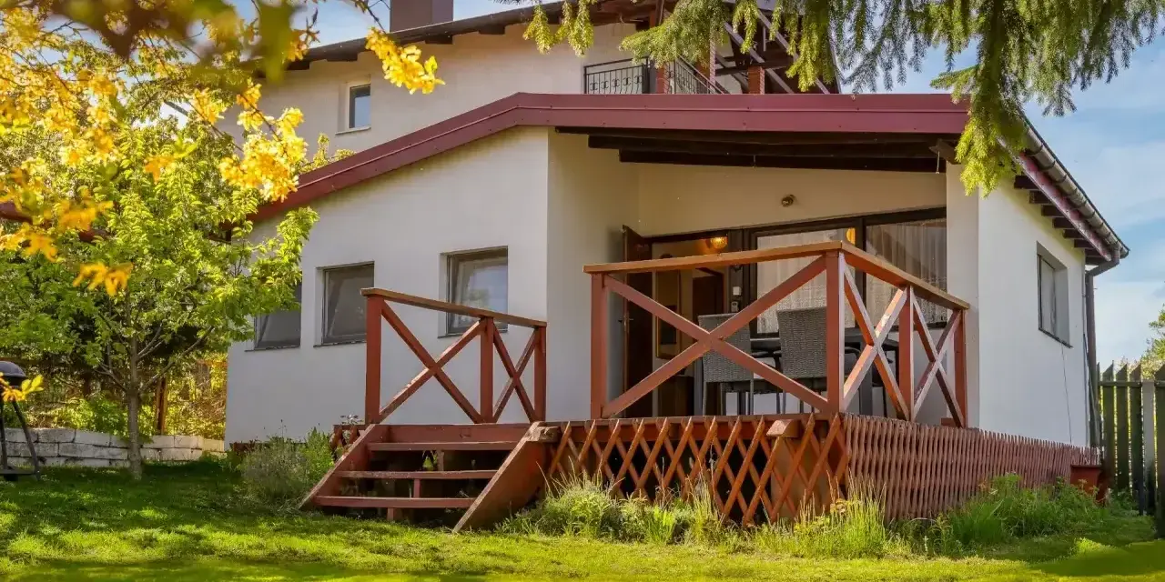 Bungalow z drewnianym tarasem i schodkami, otoczony zielonym ogrodem i kwitnącym drzewem.