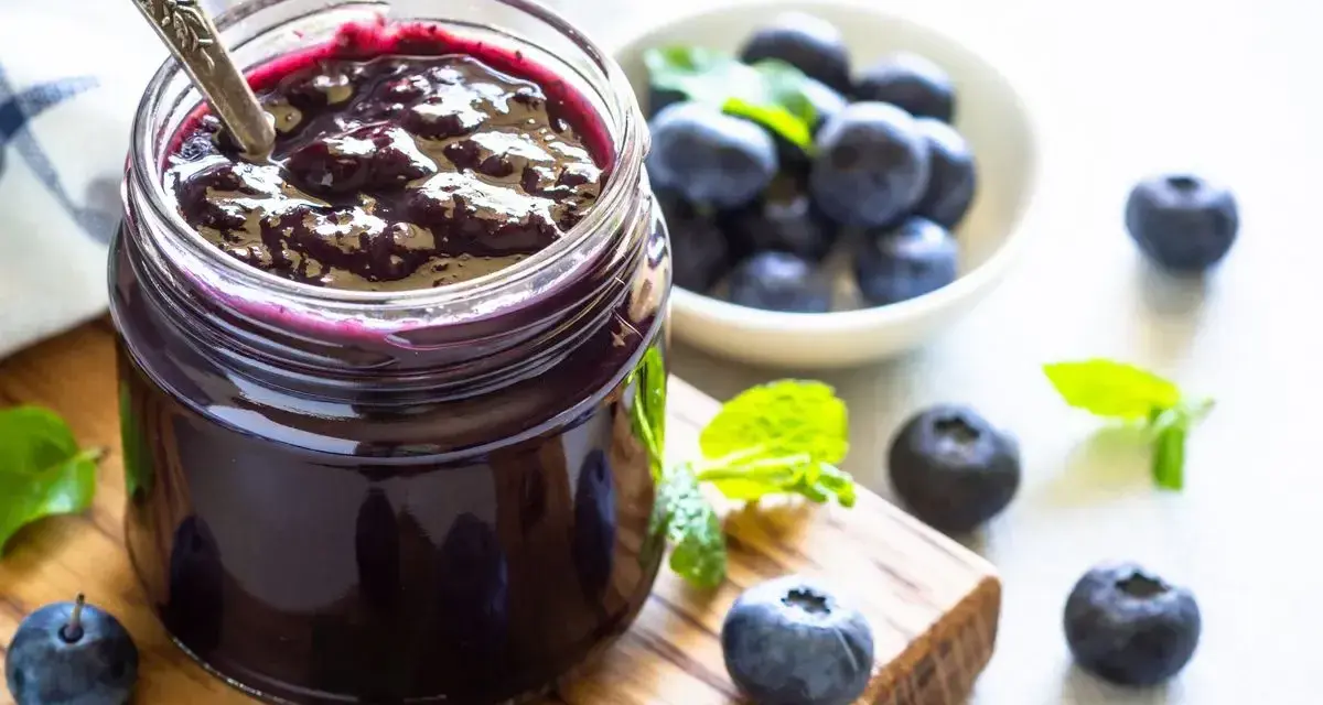 Vasetto di marmellata di mirtilli fatto in casa, con mirtilli freschi e foglie di menta su un tagliere di legno.