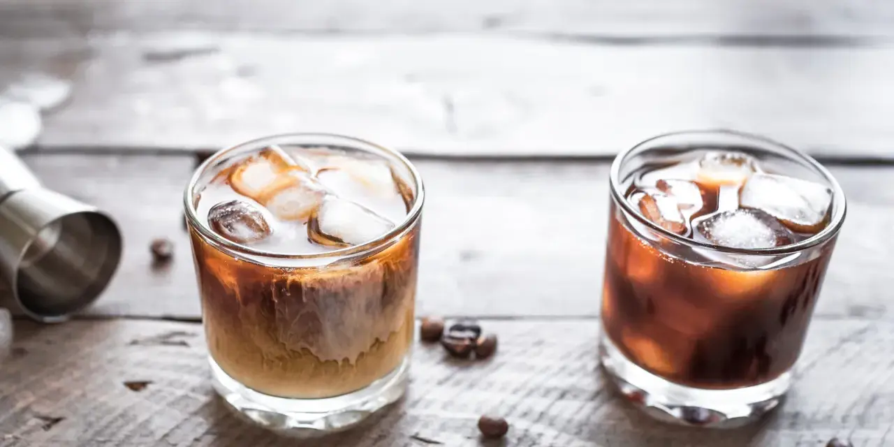 Dwa drinki na drewnianym stole: jeden to kremowy White Russian, drugi to ciemny Black Russian.