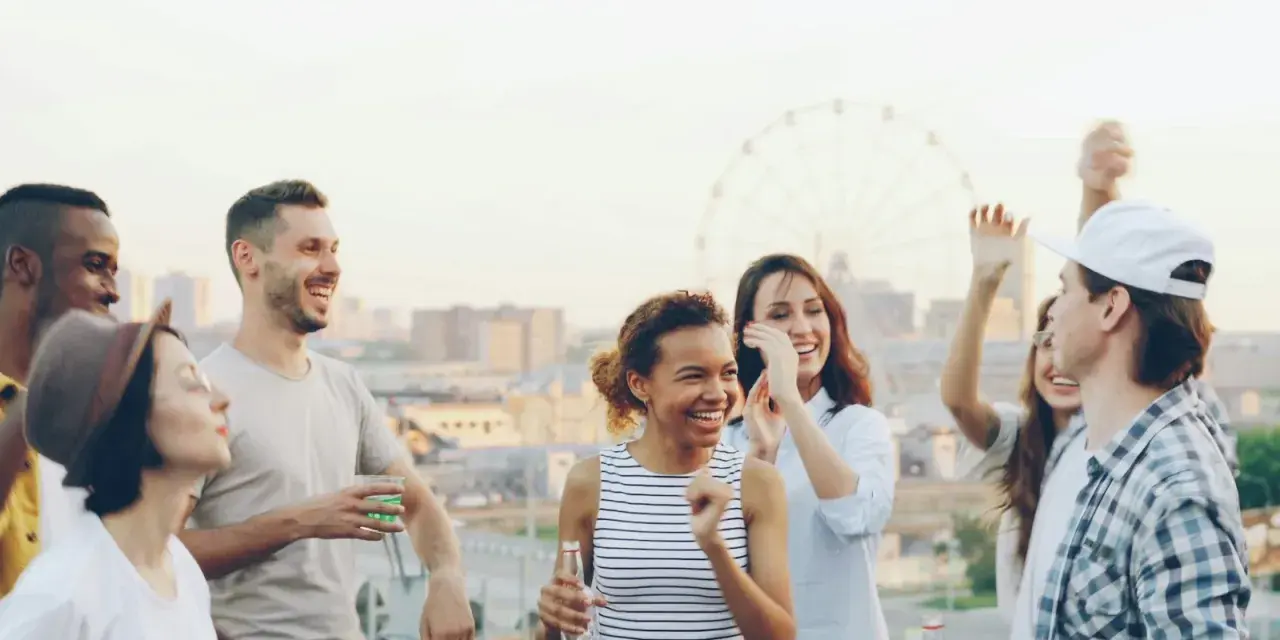 Un groupe d'amis souriants et dansant sur un toit, avec une grande roue en arrière-plan.