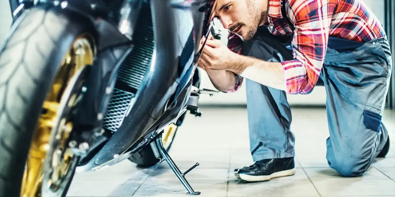 Mechanik w kratkę wykonuje przegląd motocykla, pochylając się nad jego przednią częścią.
