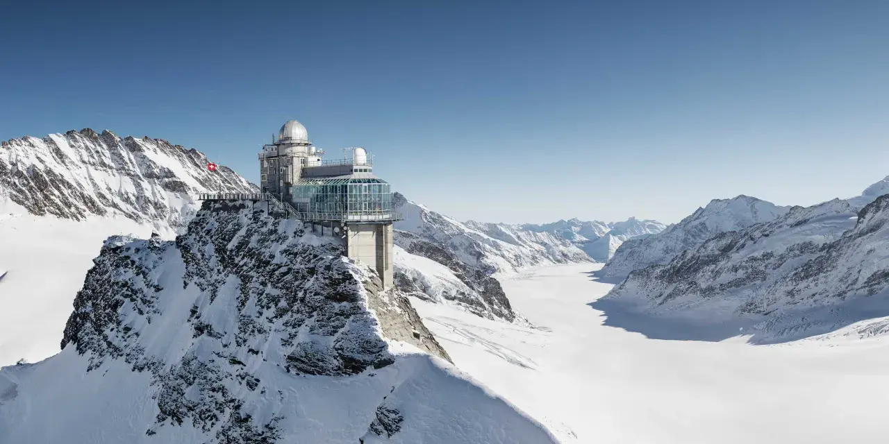 A modern building with glass walls and domes sits atop a snowy mountain, offering breathtaking views of the surrounding peaks and a vast glacier. This is the top of Europe, a place of incredible altitude.