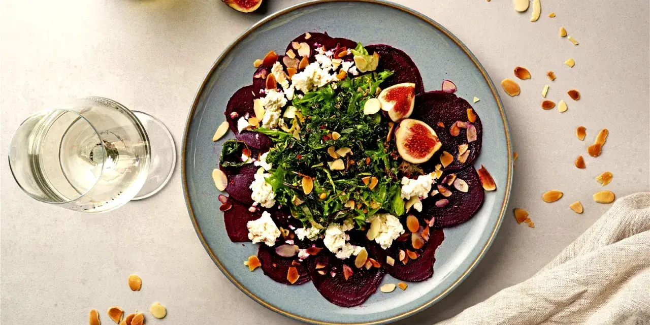 Rote Bete Carpaccio mit Feigen, Ziegenkäse und Mandelsplittern auf einem blauen Teller, daneben ein Glas Weißwein.