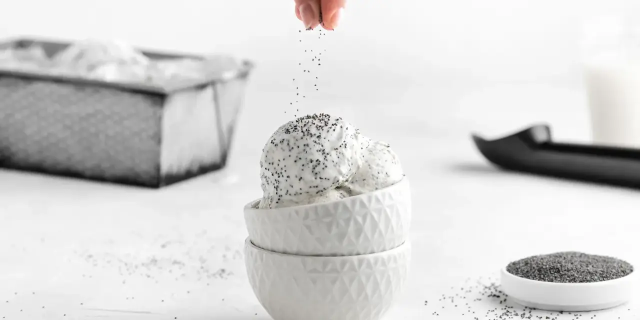 Eine Hand streut Mohn über cremiges Mohn Eis in weißen Schalen. Ein Löffel und eine Schale Mohnsamen liegen daneben.