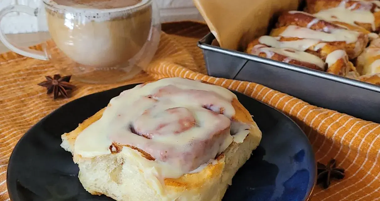 Pyszne cynamonki przepis na talerzu, obok kubek kawy i blaszka z innymi słodkościami. Idealne na jesienne popołudnie.