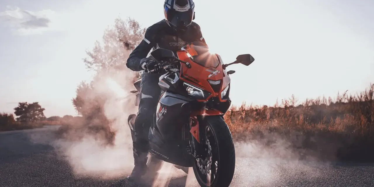Motocyklista w kasku i kombinezonie na pomarańczowym motocyklu Barton Blade, z opon dymiących na drodze.
