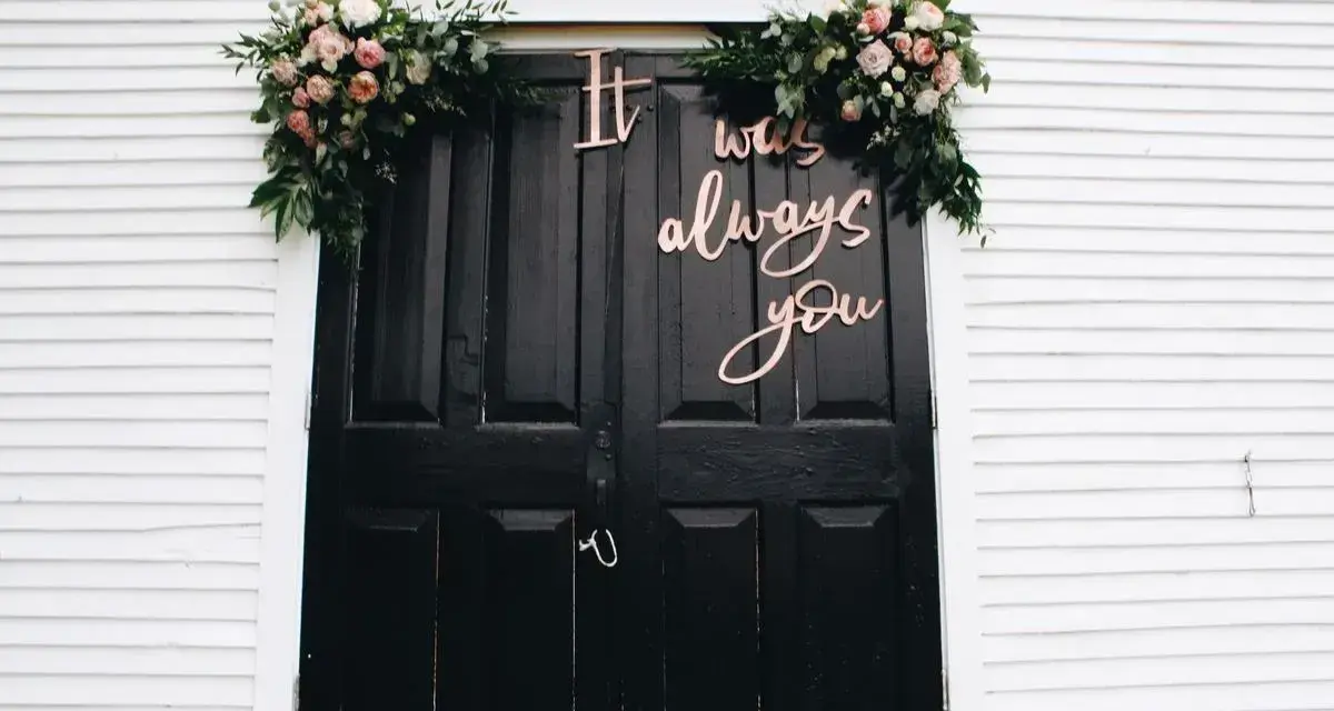 Przed domem dekoracja domu na wesele: czarne drzwi ozdobione kwiatami i napisem "It was always you".