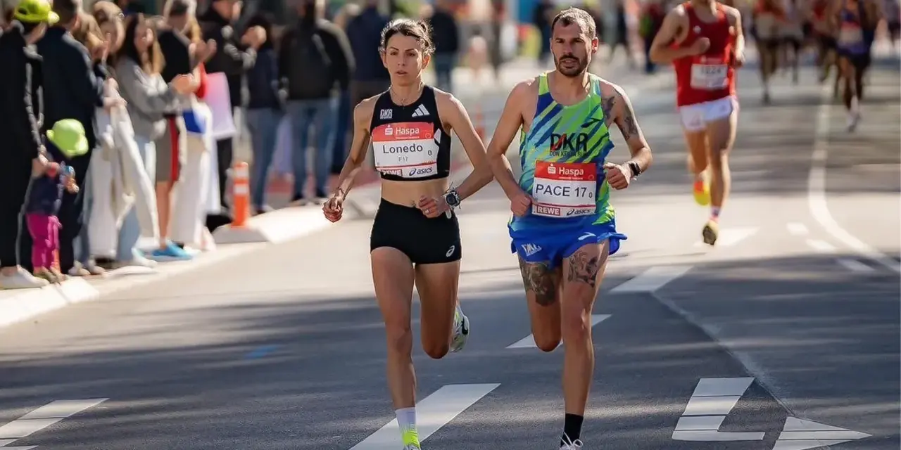 Atleti si preparano a una maratona, correndo fianco a fianco. La folla incita lungo il percorso.
