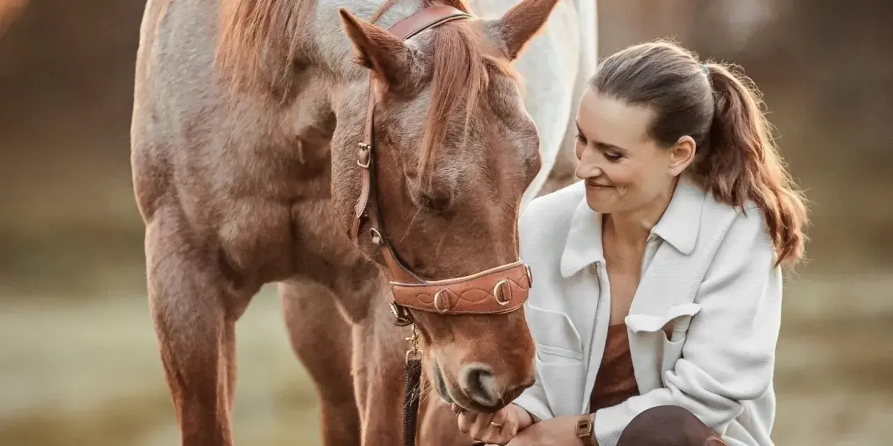 Eine Frau kniet neben einem braunen Pferd und streichelt es sanft. Selbst ein hektisches Pferd kann durch ruhiges Reiten und liebevolle Zuwendung gezähmt werden.