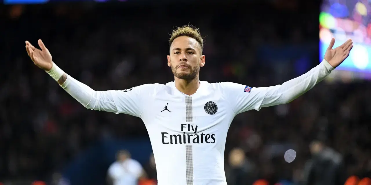 Neymar w białej koszulce PSG, z charakterystyczną neymar fryzurą, unosi ręce w geście radości na tle rozmazanych świateł stadionu.
