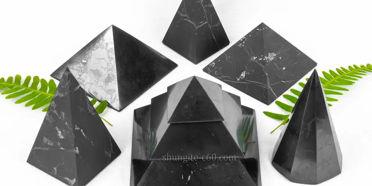 Seis pirámides de piedra shungita, algunas con vetas blancas, dispuestas sobre un fondo blanco con hojas verdes.