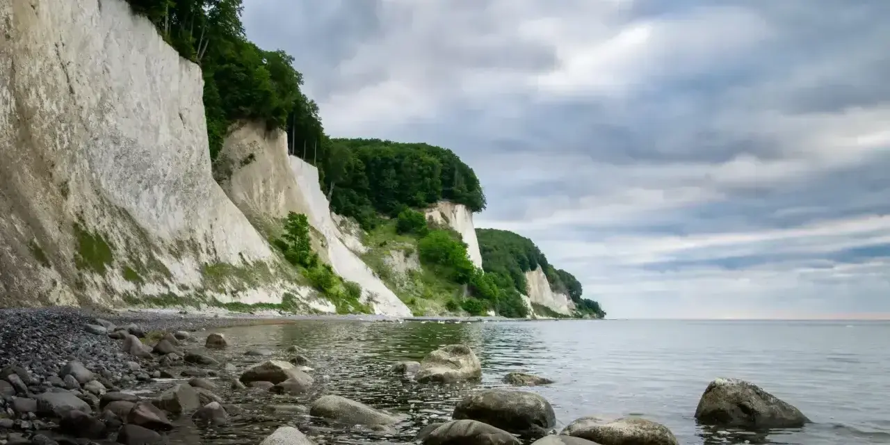 Majestatyczne, białe klify Rugii wznoszą się nad kamienistą plażą i spokojnym morzem. Zielone drzewa porastają szczyty, a chmury tworzą dramatyczne tło.
