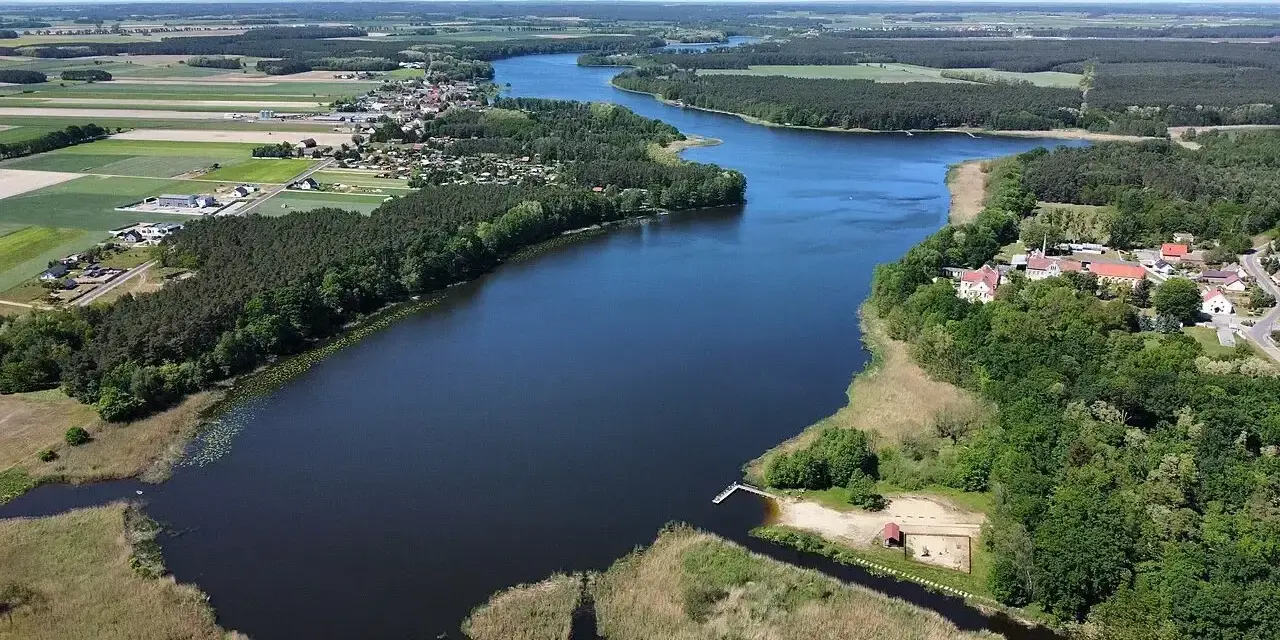Malownicze jezioro Wojnowskie otoczone lasami i polami. W oddali widać zabudowania wsi, a na brzegu plażę z pomostem.