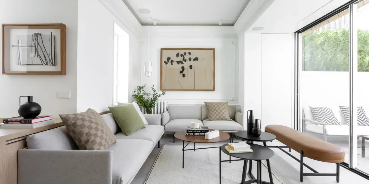 Salón moderno con sofás grises, mesas bajas negras y marrones, y un banco de cuero. Grandes ventanales conectan el interior con la terraza, creando un espacio fluido para unir terraza y salón.