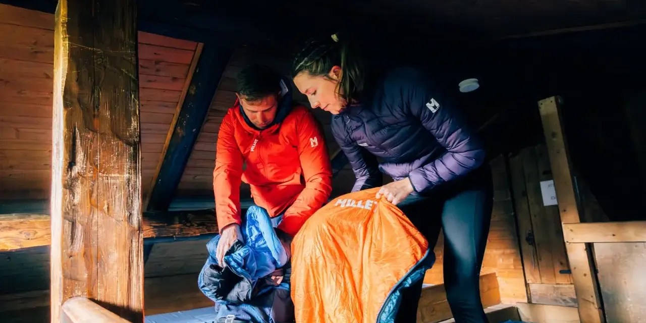 Préparation pour une nuit en refuge. Un homme en veste rouge et une femme en veste violette déballent des sacs de couchage dans une cabane en bois.