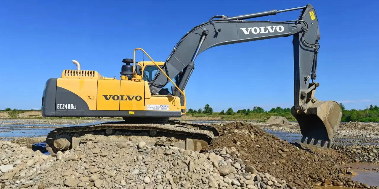 Żółta koparka Volvo EC240B LC pracuje na kamienistym brzegu rzeki. Zastanawiasz się, ile kosztuje godzina pracy koparki?