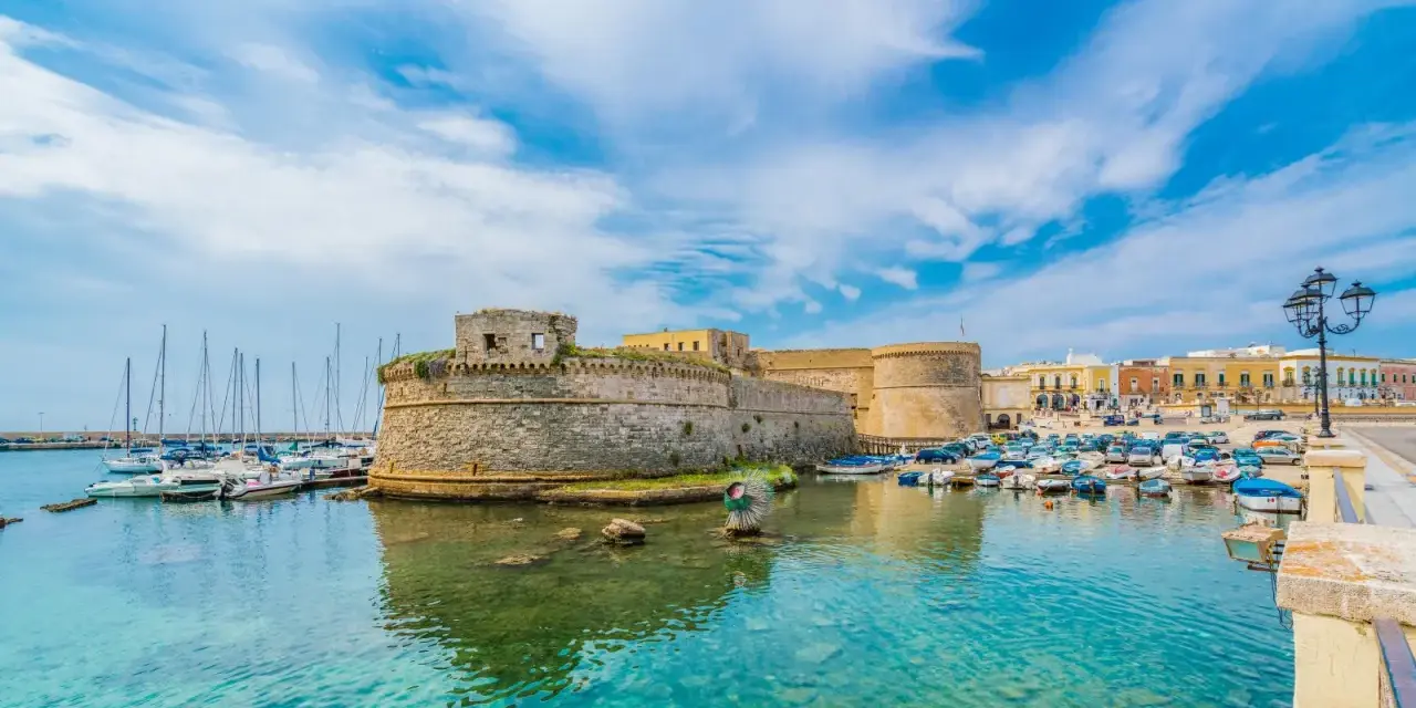 A stone castle overlooks a harbor filled with boats and yachts under a bright blue sky.