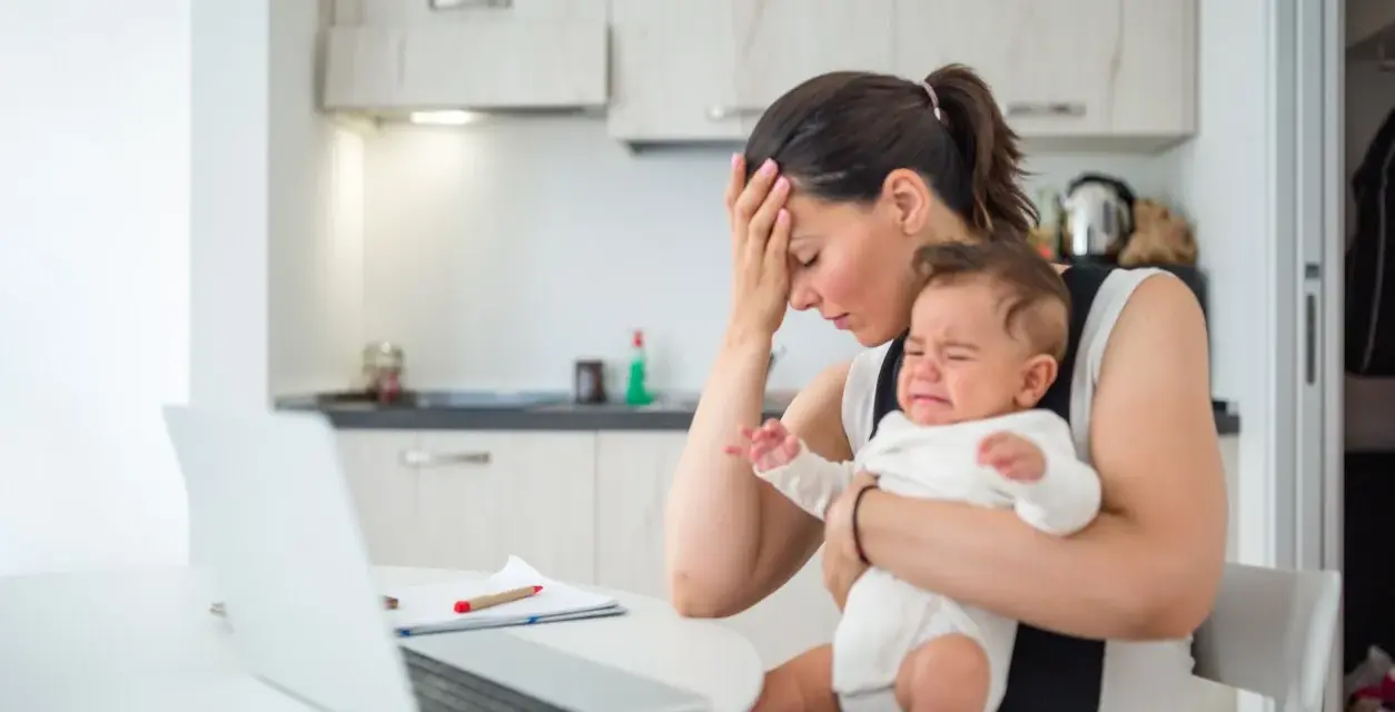 Zmęczona mama z płaczącym niemowlakiem na rękach, próbuje pracować przy laptopie. Czuje przebodźcowanie niemowlaka i własne.