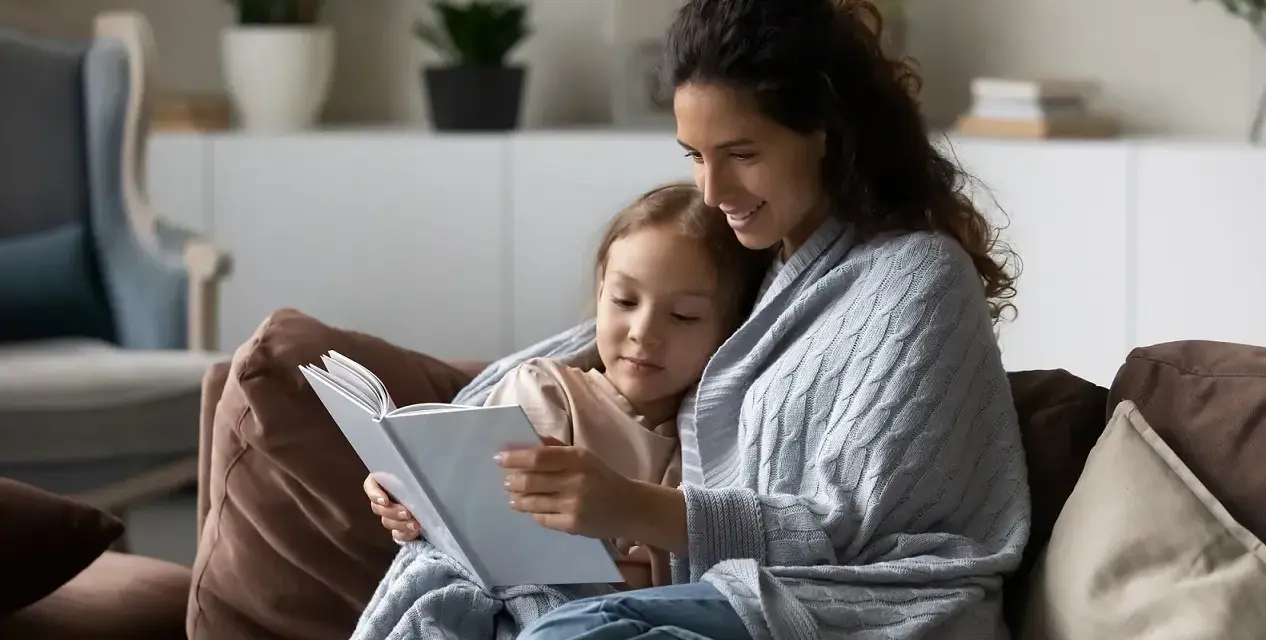Mama czyta dziecku bajkę, otuleni kocem. Widać, ile jest opieki na dziecko w tej chwili.