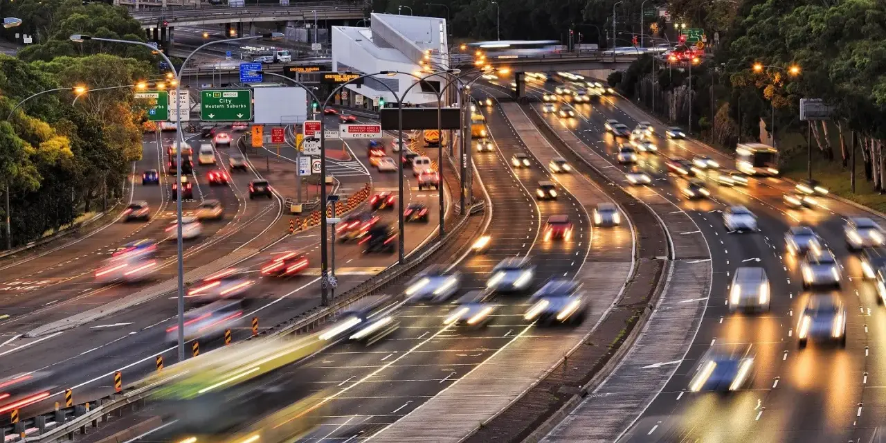 Traffico intenso su un'autostrada urbana al tramonto. Le luci dei veicoli creano scie luminose, con un **segnale 105** che indica la direzione.