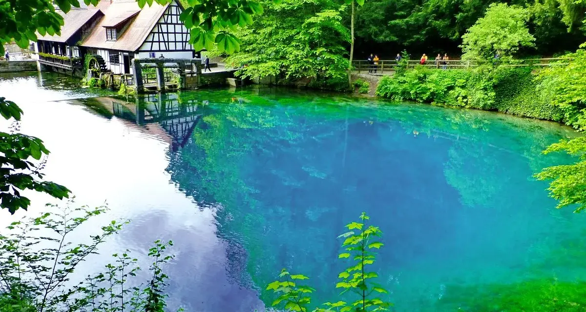 Der Blautopf See lädt zum Schwimmen ein. Ein historisches Fachwerkhaus mit Wasserrad spiegelt sich im kristallklaren, blauen Wasser.
