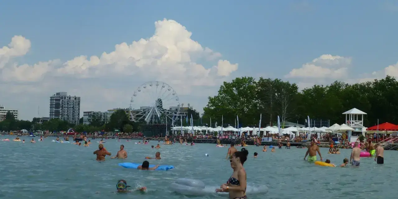 Słoneczna balaton plaża tętni życiem. Ludzie kąpią się, pływają na materacach i odpoczywają pod parasolami. W tle widać diabelski młyn.