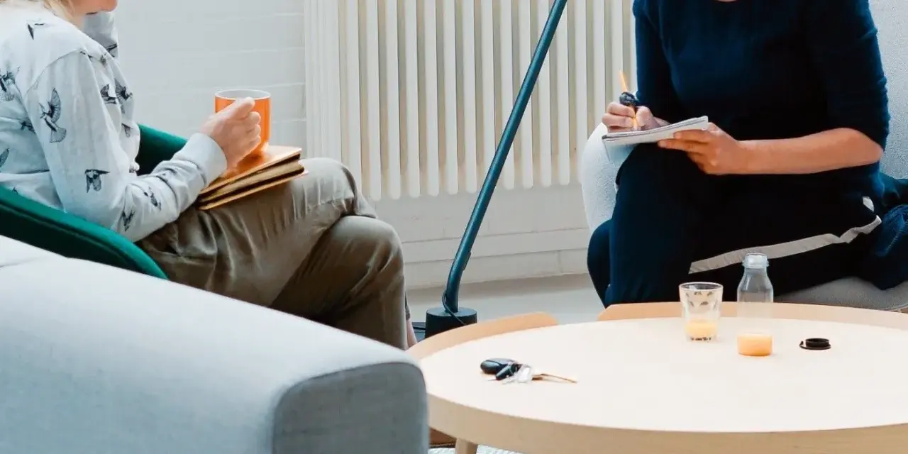 Dwie osoby siedzą w fotelach, jedna trzyma kubek, druga pisze w notatniku.