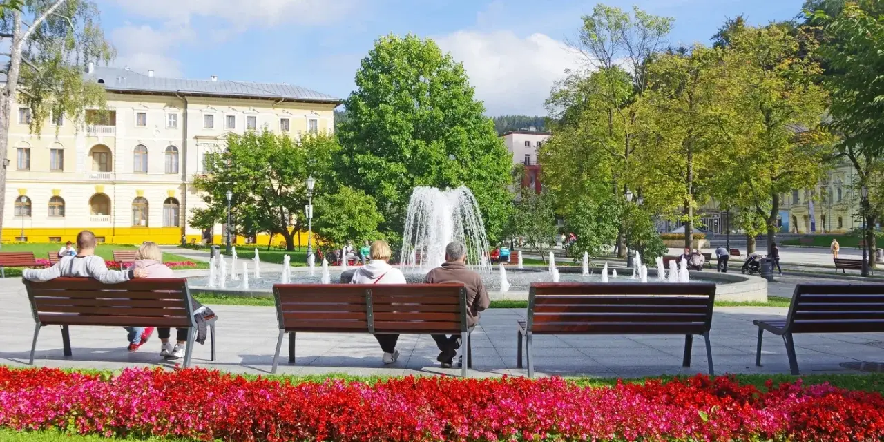 Park z fontanną i ludźmi siedzącymi na ławkach, w tle budynek.