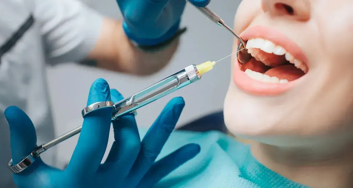 Dentysta podaje znieczulenie, by leki przeciwbólowe zadziałały przed zabiegiem. Pacjentka ma otwarte usta, a w nich widać narzędzia stomatologiczne.