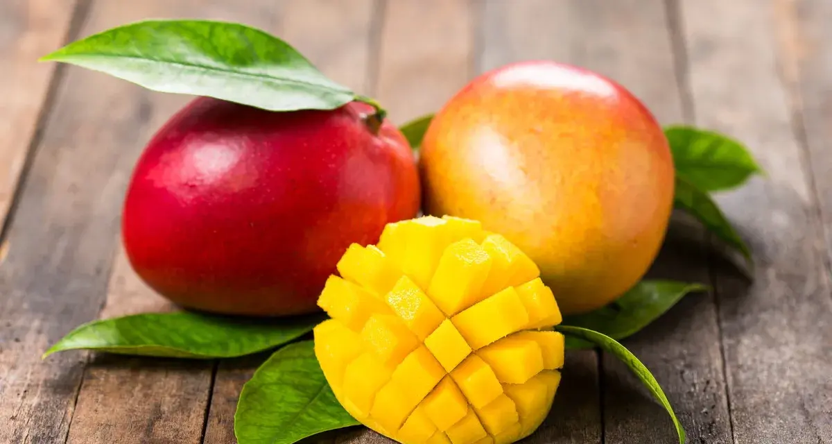 Dwa soczyste mango, jedno czerwone, drugie pomarańczowe, leżą na drewnianym blacie. Obok nich pokrojone w kostkę, złociste wnętrze owocu. To przypomina o tropikalnych krajach, skąd pochodzi mango.