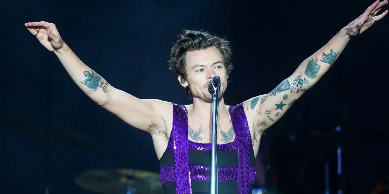 Harry Styles w błyszczącym fioletowym topie śpiewa na koncercie, unosząc ramiona. Jego ramiona pokryte są tatuażami.