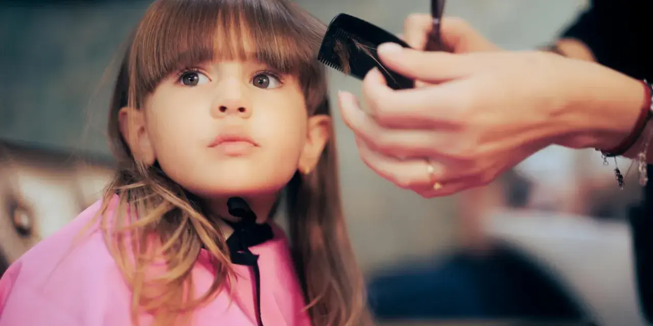 Ein **Kind** bekommt einen neuen **Pony schneiden** beim Friseur. Die kleine Dame trägt einen pinken Umhang.