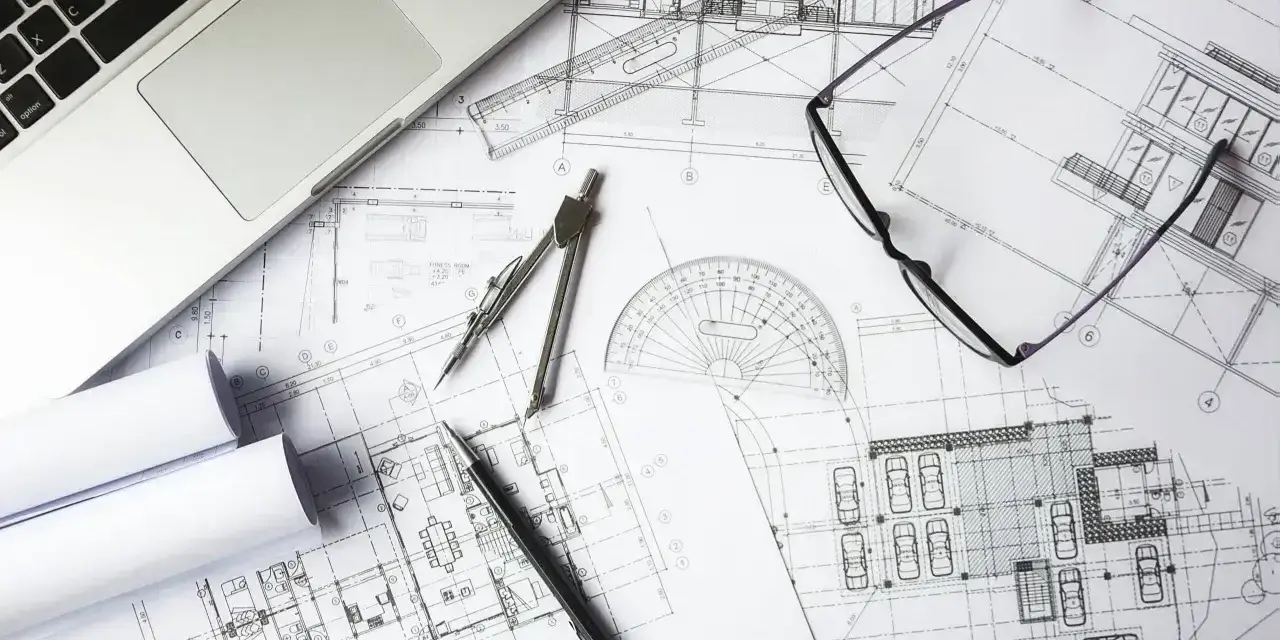 Na biurku architekta leżą plany budowlane, laptop, okulary i cyrkiel. Trwają prace nad przedmiarem robót.