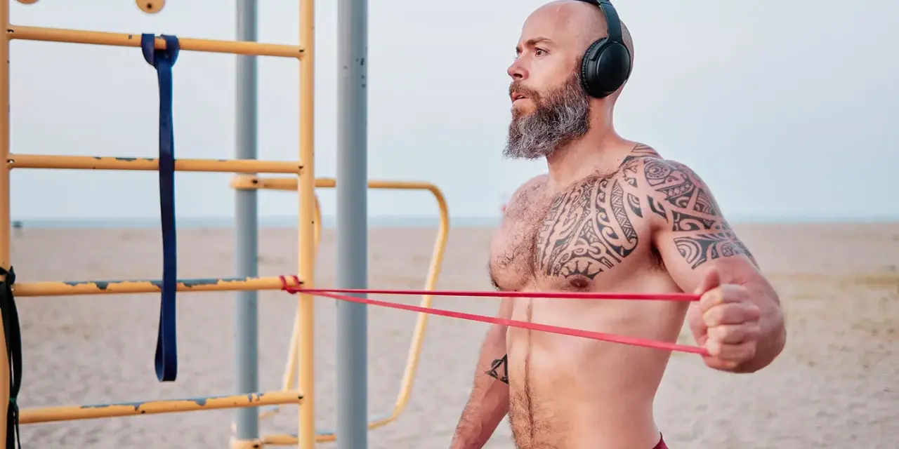 Man med tatueringar och skägg tränar med gummisnoddar vid ett utegym på stranden.