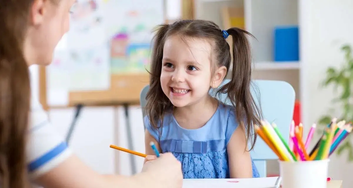 Uśmiechnięta dziewczynka z kitkami uczy się rysować. To ważny element jej obowiązku szkolnego, wspierany przez nauczycielkę.
