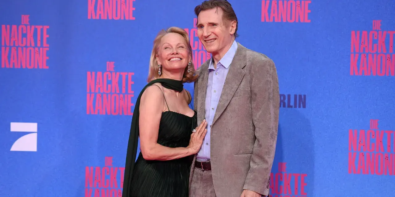 Liam Neeson e Pamela Anderson sorridono sul tappeto blu, con lo sfondo di "Die Nackte Kanone".