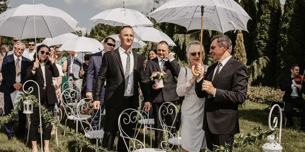 Wesele w kwietniu pod białymi parasolami. Goście w eleganckich strojach, wśród nich pan młody z bukiecikiem.