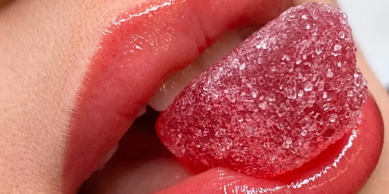 Labbra carnose e lucide, con un effetto volume dato da un tatuaggio labbra, assaporano una caramella rossa zuccherina.